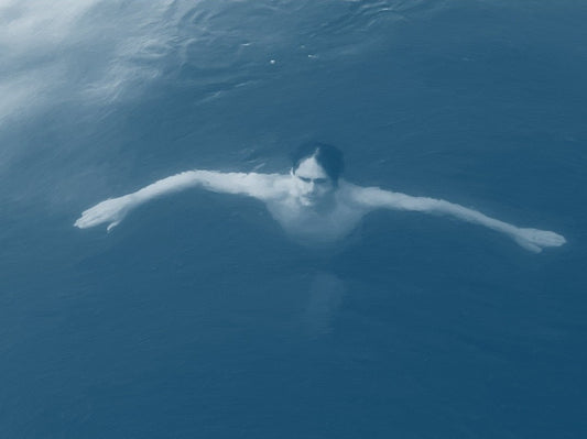 Das Kunstwerk "Christianisierung Brandenburgs" des Künstlers nostrokesbitte zeigt ein digitales Gemälde im fotorealistischen Stil. Es ist ein Porträt, das eine Person von oben im Wasser liegend darstellt. Die Person blickt direkt auf den Betrachter und hat die Arme ausgebreitet. Das Bild ist in kühlen, bläulichen Farbtönen gehalten, die die Wasseroberfläche darstellen. Das Werk wurde auf Leinwand gedruckt und ist in einer limitierten Edition über die Online-Galerie FEMARTSPACE erhältlich.