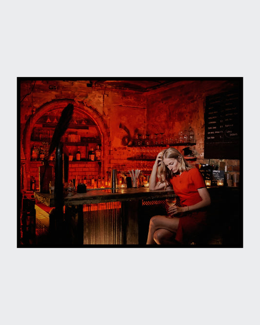 Das fotorealistische Digitalgemälde "Chablis bei Schlamassel" von nostrokesbitte zeigt eine blonde Frau im roten Kleid, die melancholisch an einer rot beleuchteten Bar sitzt und ein Glas Wein hält. Das Werk ist in limitierter Auflage bei FEMARTSPACE erhältlich.