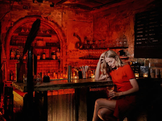 Das fotorealistische Digitalgemälde "Chablis bei Schlamassel" von nostrokesbitte zeigt eine blonde Frau im roten Kleid, die melancholisch an einer rot beleuchteten Bar sitzt und ein Glas Wein hält. Das Werk ist in limitierter Auflage bei FEMARTSPACE erhältlich.