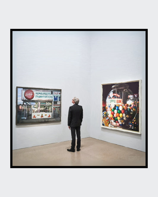 Das fotorealistische Digitalgemälde "Berlin" aus der Serie "Rueckenfiguren" von nostrokes zeigt einen Mann in einem schwarzen Anzug, der von hinten in einer Kunstgalerie steht. Er betrachtet zwei große, farbige Gemälde im Stil des Fotorealismus an einer weißen Wand. Das quadratische Werk ist in einer limitierten Edition auf Leinwand gedruckt und in der Online-Galerie FEMARTSPACE erhältlich.