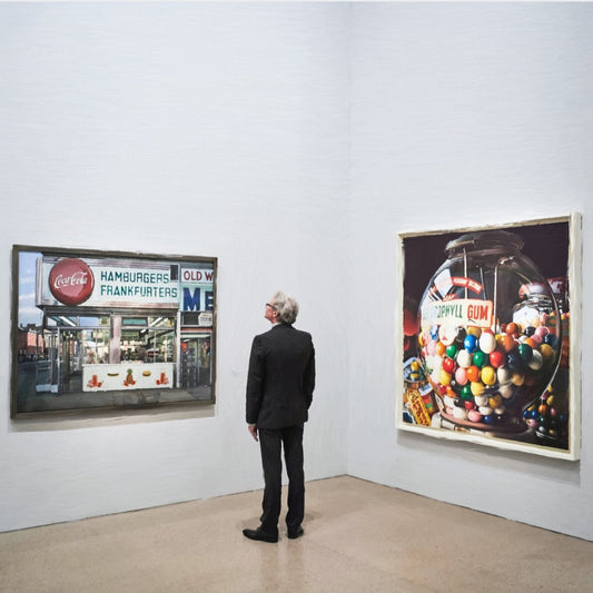 Das fotorealistische Digitalgemälde "Berlin" aus der Serie "Rueckenfiguren" von nostrokes zeigt einen Mann in einem schwarzen Anzug, der von hinten in einer Kunstgalerie steht. Er betrachtet zwei große, farbige Gemälde im Stil des Fotorealismus an einer weißen Wand. Das quadratische Werk ist in einer limitierten Edition auf Leinwand gedruckt und in der Online-Galerie FEMARTSPACE erhältlich.