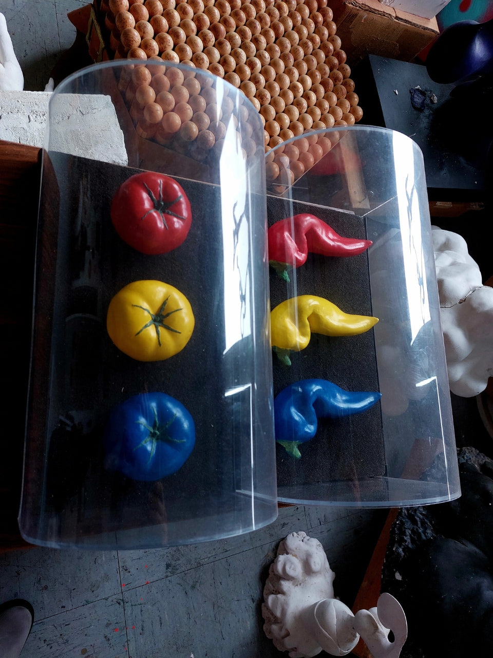 Skulptur von Amos Plaut (FEMARTSPACE): Zwei zylinderförmige, transparente Kunststoffbehälter stehen nebeneinander auf einem grauen, unebenen Untergrund. In jedem Behälter befinden sich drei stilisierte, organisch geformte Objekte in den Farben Rot, Gelb und Blau. Die Objekte im linken Behälter sind kompakter und ähneln stilisierten Tomaten. Die Objekte im rechten Behälter sind länglicher und gewundener, mit einer tropfenartigen Form. Im Hintergrund sind unscharf weitere Ateliergegenstände zu erkennen.