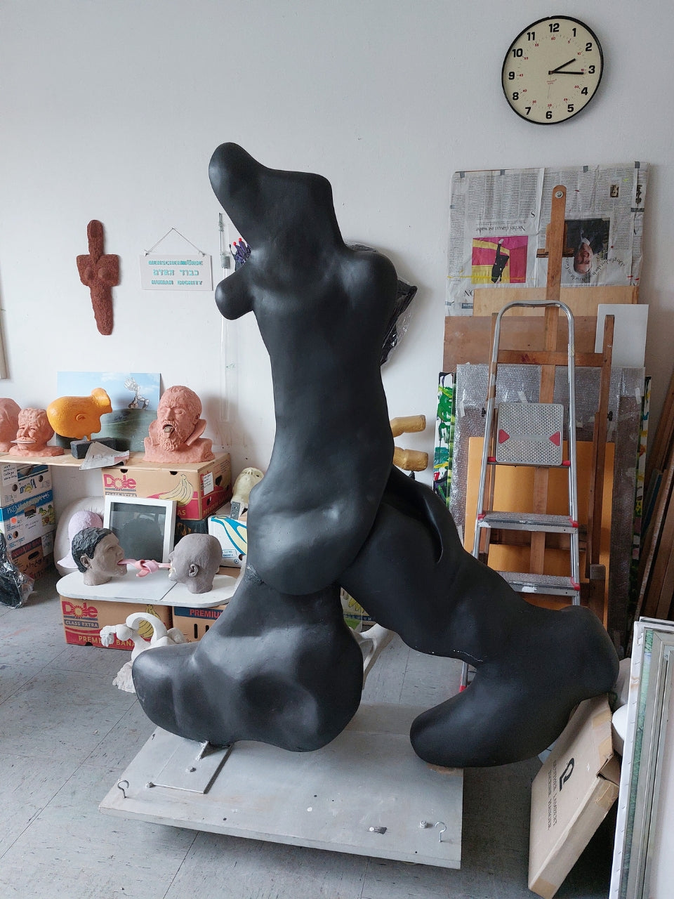 Skulptur von Amos Plaut (FEMARTSPACE): Eine große, organisch geformte Skulptur aus dunklem Material, möglicherweise Ton oder Gips, dominiert den Raum eines Ateliers. Die Skulptur erinnert an einen stilisierten, liegenden oder sich windenden Körper mit abgerundeten Formen und Ausstülpungen. Sie steht auf einer hellen, rechteckigen Basis. Im Hintergrund ist ein weiß gestrichener Raum mit verschiedenen weiteren Kunstwerken, Skulpturen auf Regalen, einer Leiter, Zeitungen an der Wand & einer runden Uhr zu sehen
