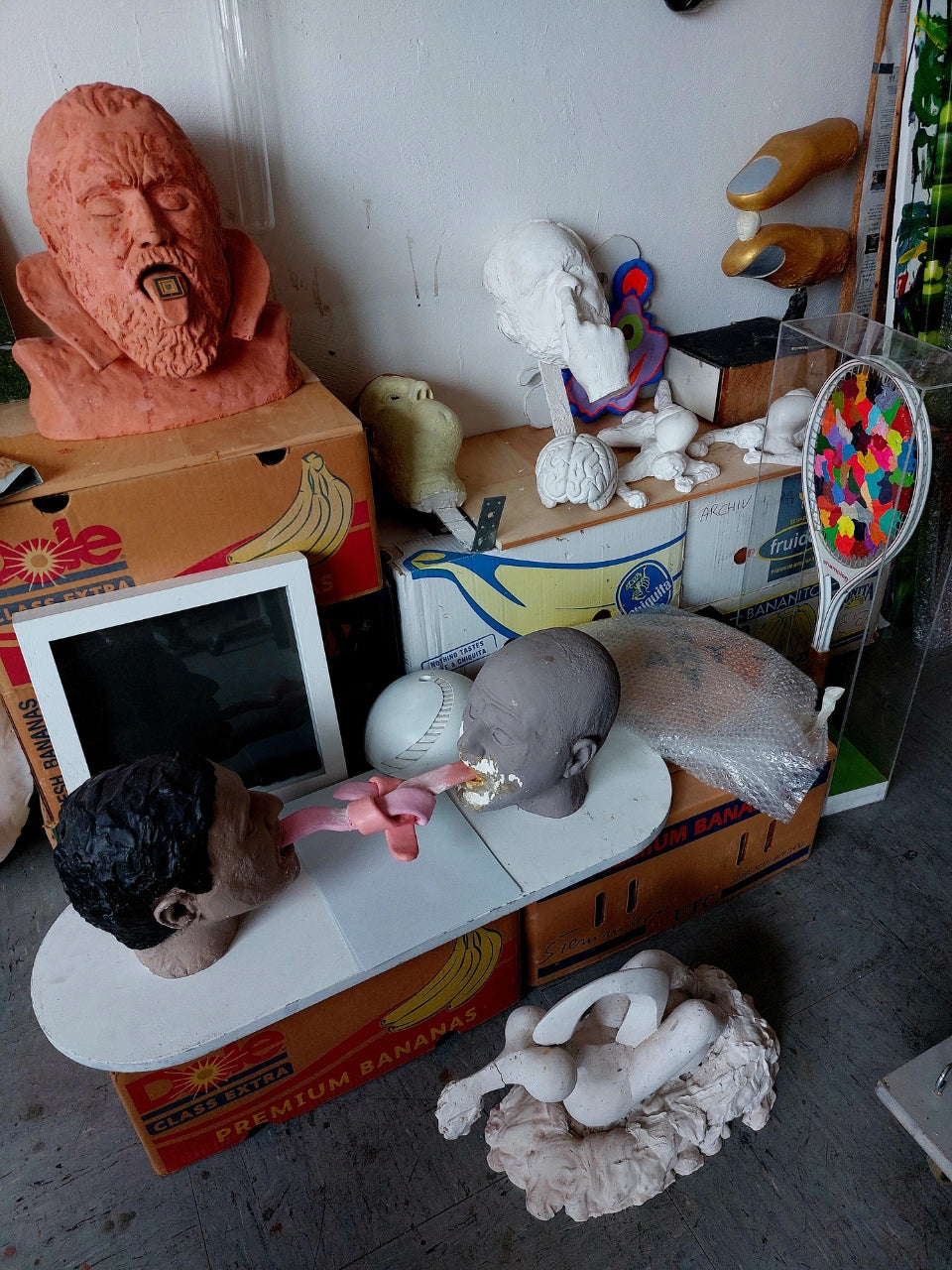 Atelier von Amos Plaut (FEMARTSPACE): Ein unaufgeräumter Arbeitsbereich voller unfertiger und fertiger Kunstwerke. Auf Kartons und einer weißen Platte liegen verschiedene Skulpturen aus unterschiedlichen Materialien wie Ton und Gips, darunter mehrere Kopfstudien mit expressiven Gesichtszügen. Ein rotbrauner, fragmentierter Kopf mit aufgerissenem Mund liegt auf einem Bananenkarton. Ein weiterer, grauer Kopf ist auf einer weißen Unterlage platziert. Im Hintergrund lehnen weitere Objekte an einer weißen Wand,