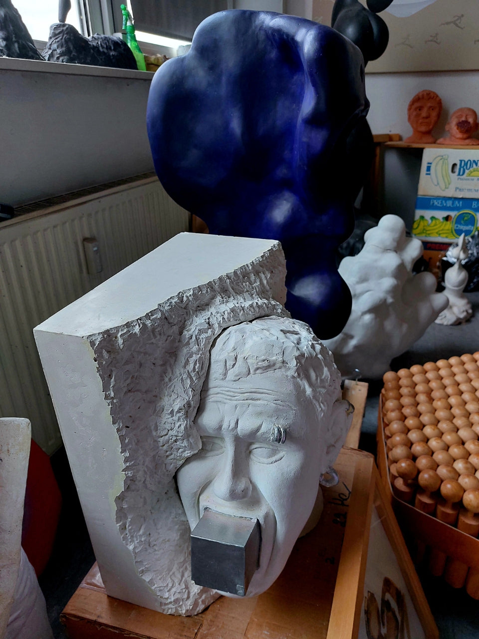 Skulptur von Amos Plaut (FEMARTSPACE): Eine weiße Gipsbüste eines Gesichtes, das in einem grob behauenen, weißen Block zu stecken scheint. Das Gesicht hat geschlossene Augen und einen nach unten gezogenen Mund, in den ein metallisch-grauer Würfel eingefügt ist. Im Hintergrund sind weitere, unscharfe Skulpturen und Ateliergegenstände zu erkennen, darunter eine große, dunkelblaue, organisch geformte Skulptur.