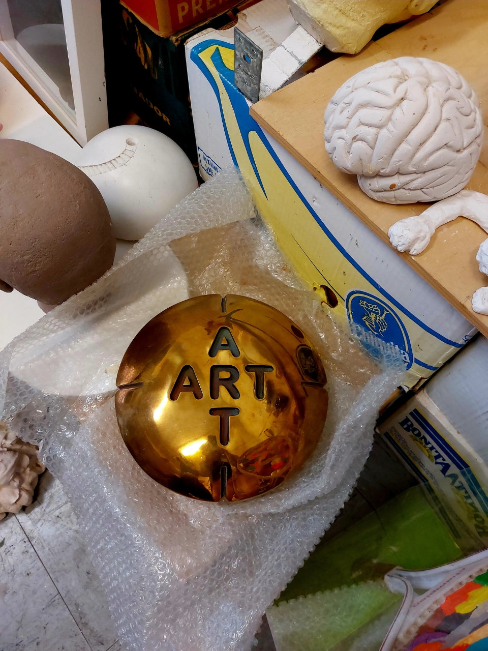 Installation 'Pille für die Kunst' von Amos Plaut (FEMARTSPACE): Eine glänzende, goldfarbene, runde Dose oder Schale liegt auf einer zerknitterten, durchsichtigen Folie. In die Oberfläche der Dose sind die Buchstaben 'ART' in einer vertikalen Anordnung eingeprägt, wobei das 'A' oben und das 'T' unten steht. Um die goldene Dose herum liegen verschiedene andere Objekte, darunter ein weißes, geformtes Objekt, das an ein Gehirn erinnert, Fragmente von Kartons mit Bananenaufdruck.