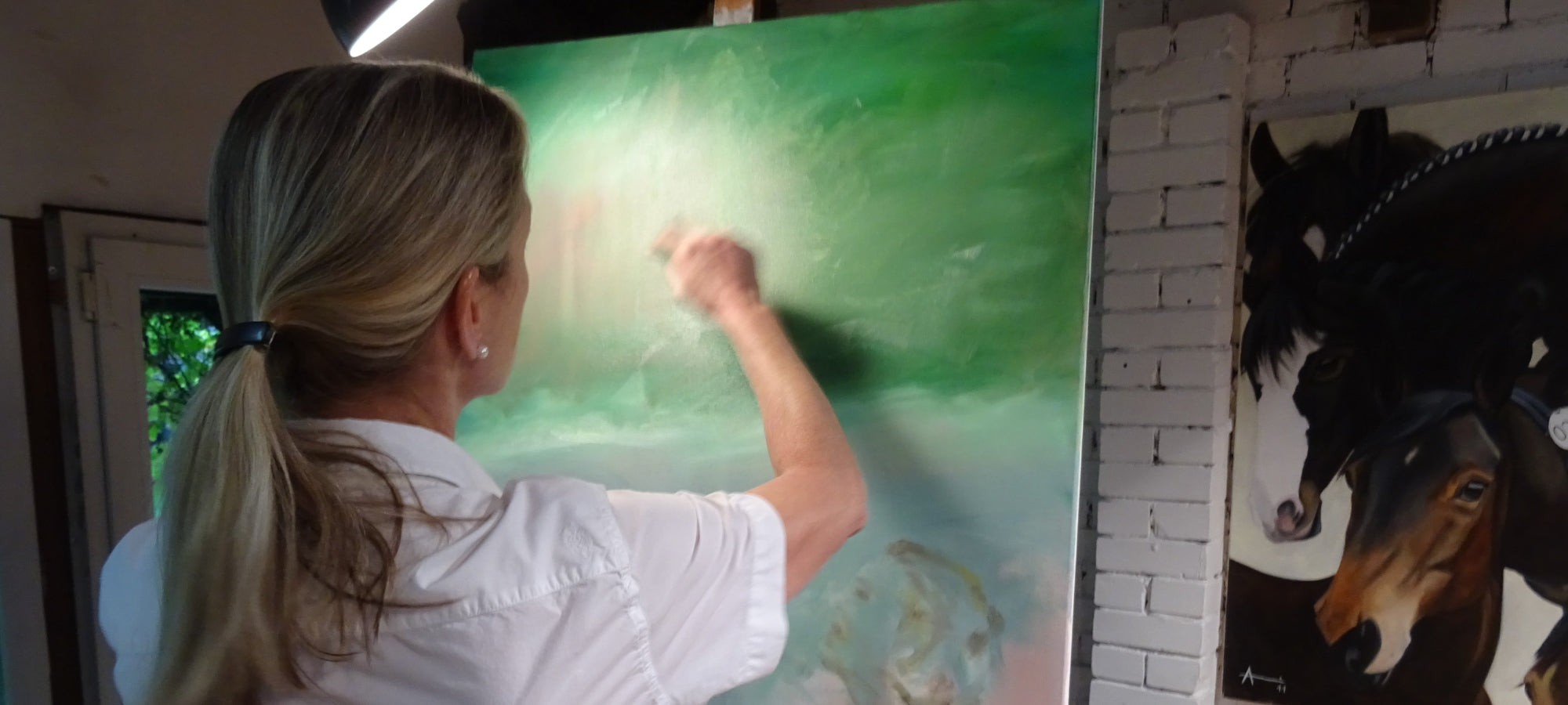 Eine blonde Frau von hinten, die vor einer grünen Leinwand steht und mit der rechten Hand malt. Links ist eine Tür zu sehen, rechts ein Teil eines anderen Gemäldes mit Pferdeköpfen. Es handelt sich um die Künstlerin Anke Dinkelbach von FemArtSPACE.