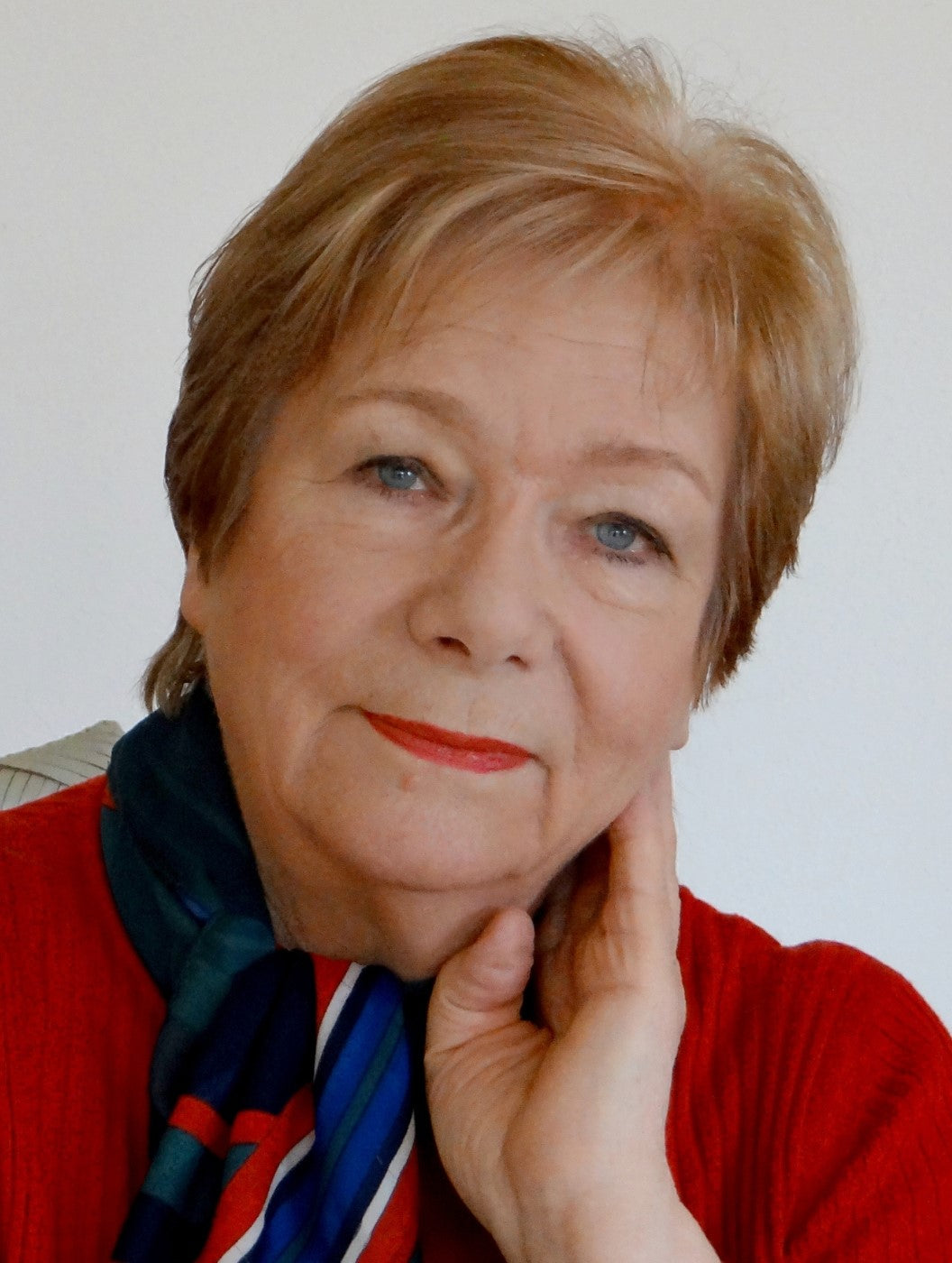 Porträt von Anke Huober, einer Künstlerin der Online-Galerie FemArtSPACE. Sie ist eine Frau mittleren Alters mit kurzen blonden Haaren und trägt einen roten Pullover und einen blau-grün gemusterten Schal. Sie stützt ihr Kinn auf ihrer rechten Hand und lächelt leicht.