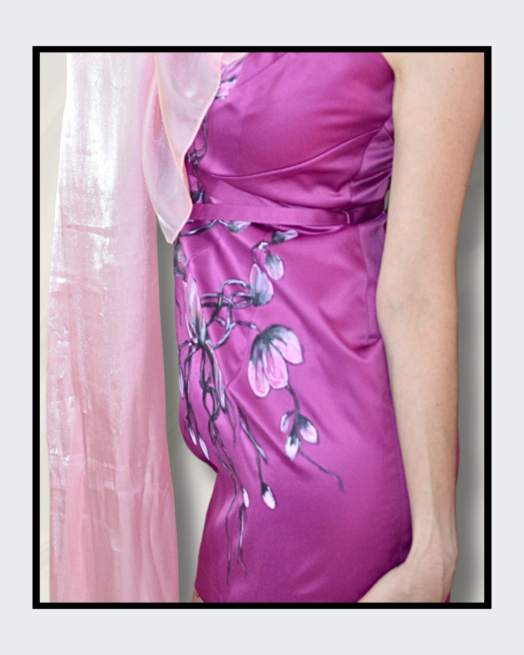 Ein elegantes, taillenbetontes Damenkleid in einem satten Purpurton. Der Stoff ist aus glänzendem Satin und mit zarten, handgemalten Blüten in verschiedenen Rosa- und Weißtönen verziert. Die Blütenranken schlängeln sich über das Kleid und verleihen ihm einen romantischen Touch. Ein zartes, durchscheinendes Tuch in einem helleren Rosaton liegt über einer Schulter und ergänzt den femininen Look. Handbemalt von Kaja Schäfer.