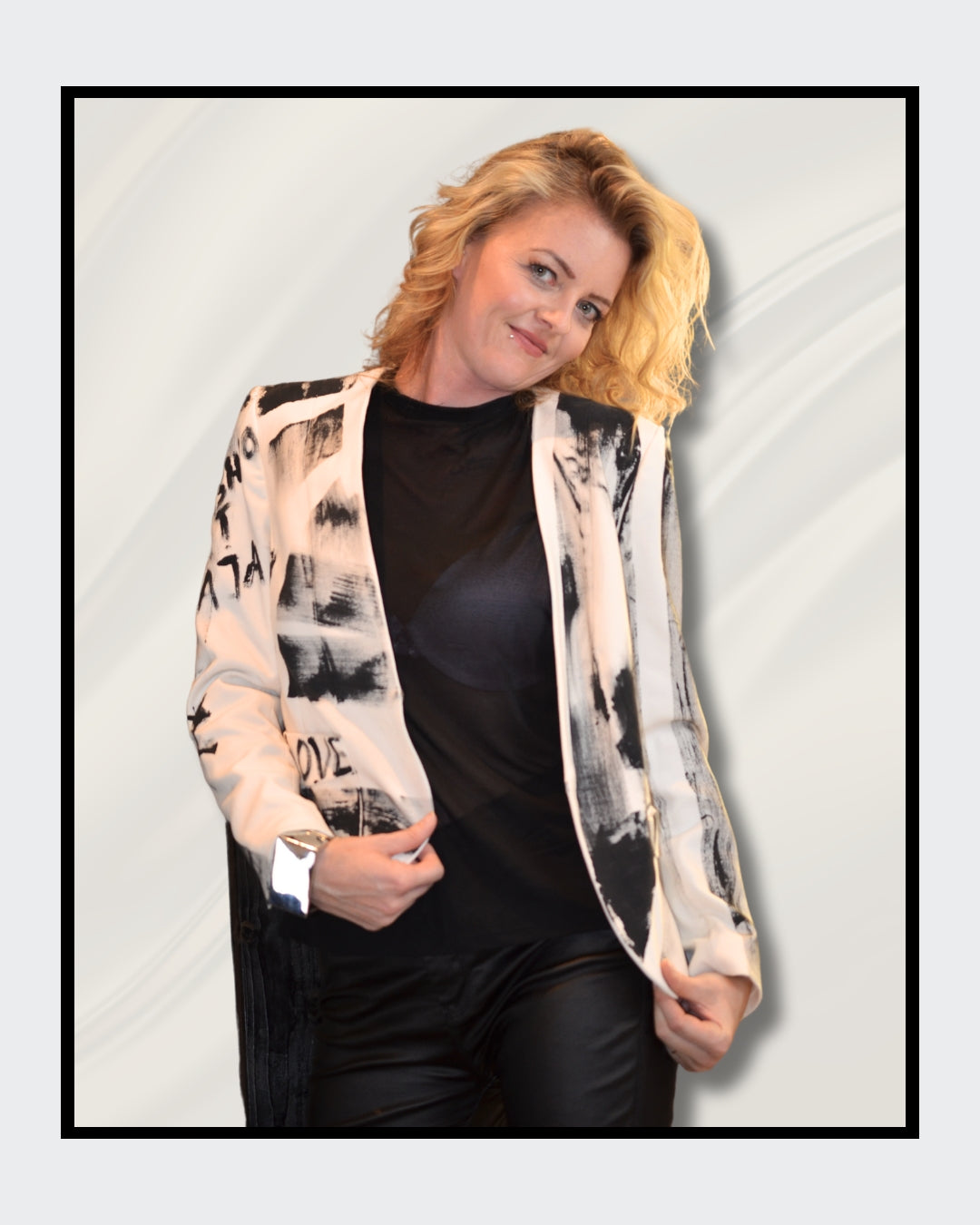 Eine blonde Frau steht vor einem cremefarbenen Hintergrund und blickt nach rechts. Sie trägt einen weißen Blazer mit schwarzen, abstrakten Mustern und Schriftzügen, darunter das Wort "LOVE". Darunter trägt sie ein schwarzes Oberteil und eine schwarze Hose. Fashionart von Kaja Schäfer, exklusiv bei FEMARTSPACE, Online-Galerie.