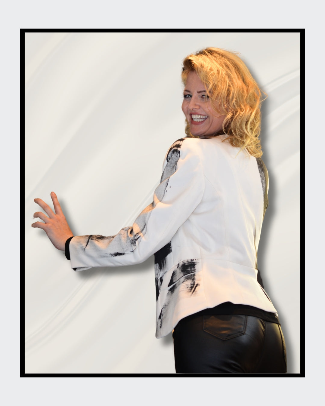 Eine blonde Frau steht vor einem cremefarbenen Hintergrund und blickt nach rechts. Sie trägt einen weißen Blazer mit schwarzen, abstrakten Mustern und Schriftzügen, darunter das Wort "LOVE". Darunter trägt sie ein schwarzes Oberteil und eine schwarze Hose. Fashionart von Kaja Schäfer, exklusiv bei FEMARTSPACE, Online-Galerie.