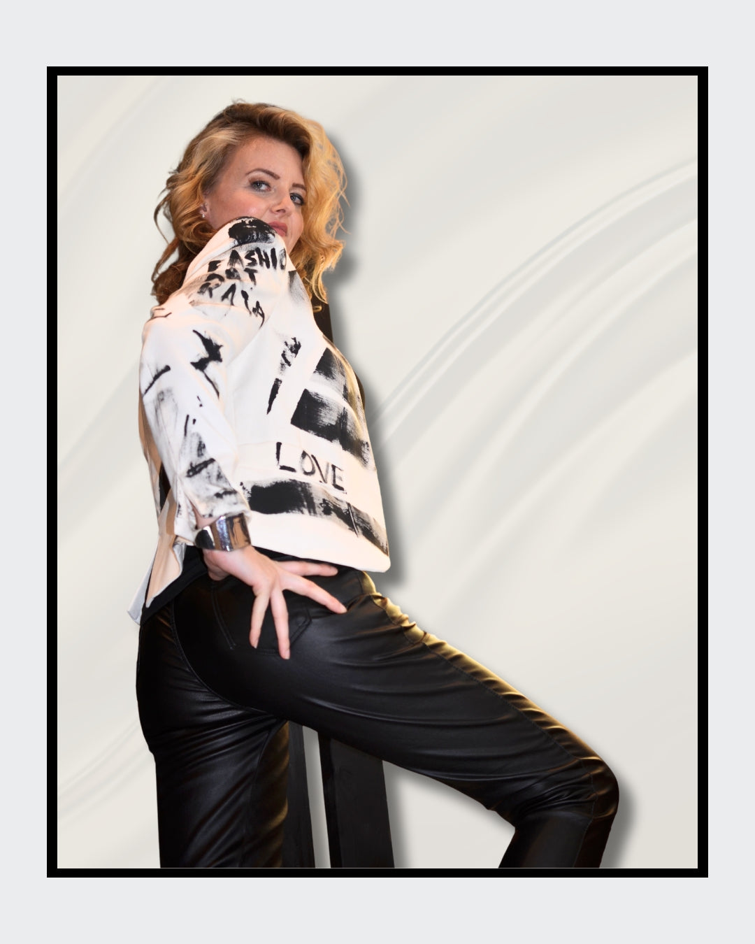 Eine blonde Frau steht vor einem cremefarbenen Hintergrund und blickt nach rechts. Sie trägt einen weißen Blazer mit schwarzen, abstrakten Mustern und Schriftzügen, darunter das Wort "LOVE". Darunter trägt sie ein schwarzes Oberteil und eine schwarze Hose. Fashionart von Kaja Schäfer, exklusiv bei FEMARTSPACE, Online-Galerie.