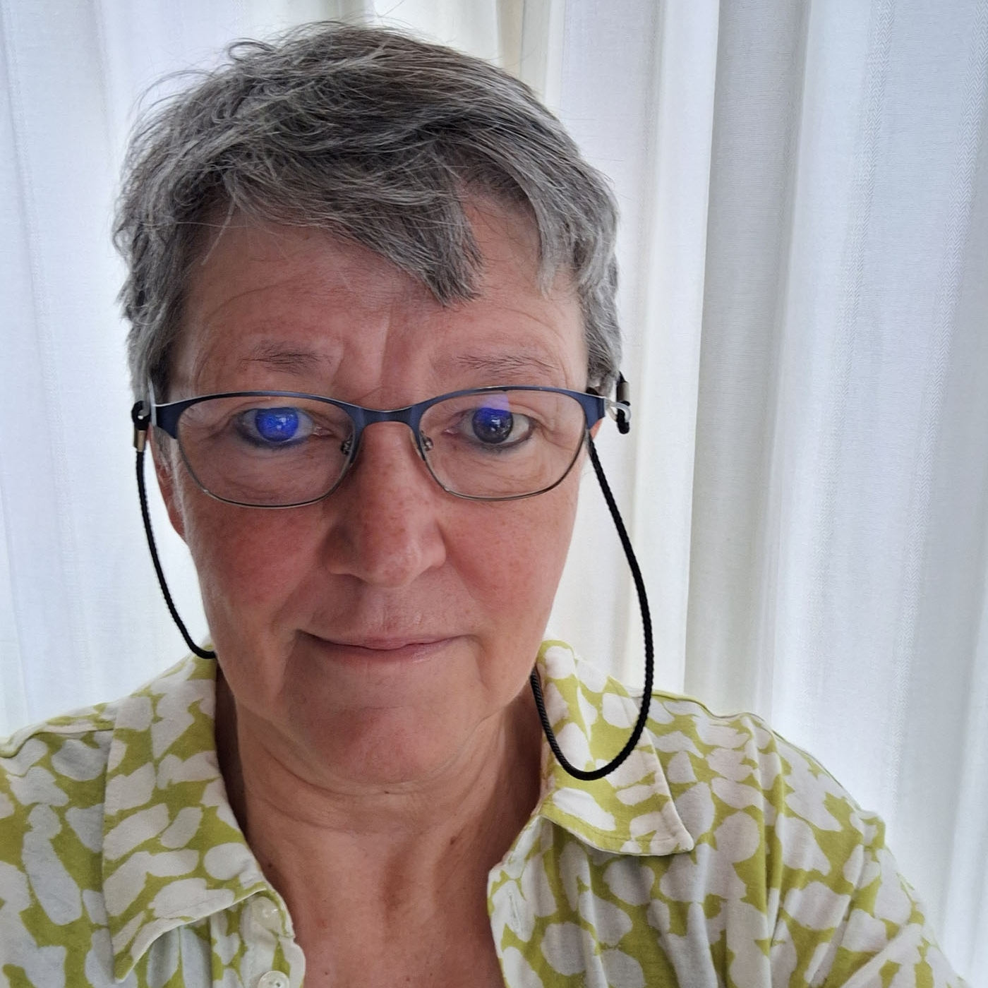 Porträtfoto der Künstlerin Monika Schiwy-Jessen mit Brille, grauem Kurzhaarschnitt und gemusterter Bluse, vor hellem Vorhang.