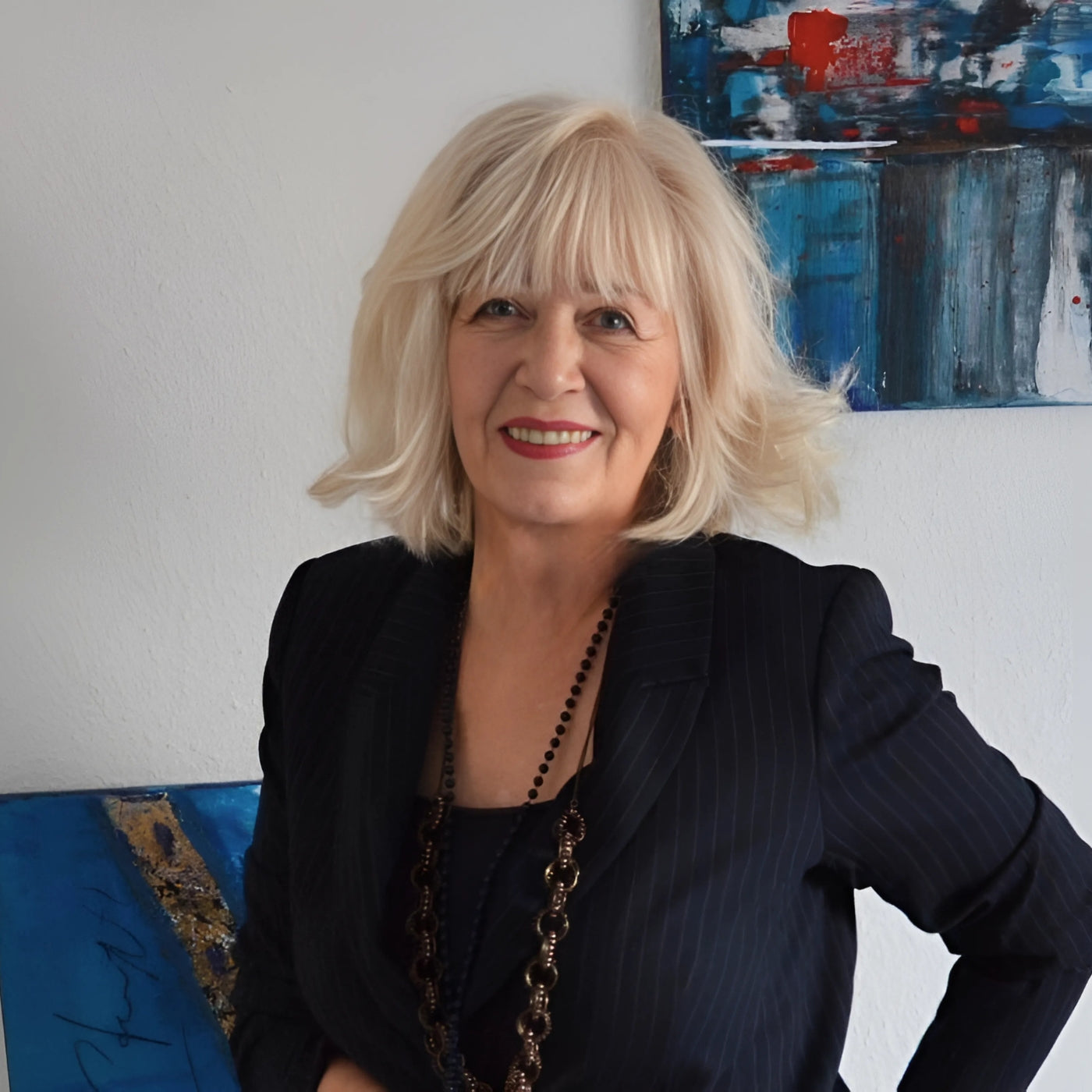 Ein Porträt der Künstlerin Olga Kühn. Sie ist eine Frau mit blonden, schulterlangen Haaren und Pony. Sie lächelt freundlich in die Kamera. Sie trägt ein dunkles, gestreiftes Jackett über einem dunklen Oberteil und eine lange, dunkle Halskette mit großen, unregelmäßigen Gliedern. Im Hintergrund sind zwei abstrakte Gemälde mit blauen, weißen und roten Farbtönen an einer hellen Wand zu sehen.