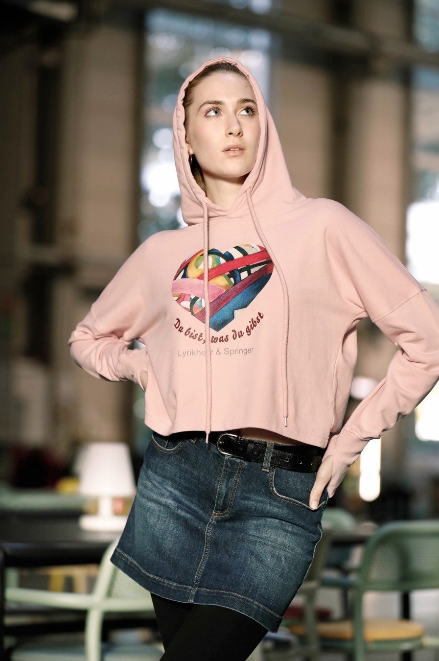 Ein kurz geschnittener, rosafarbener Hoodie mit überschnittenen Schultern. Extralange Bündchen mit Daumenloch und eine bedruckte Kunstgrafik auf der Vorderseite. Mode von Soulfashion Lyrikherz & Springer, erhältlich bei FEMARTSPACE.