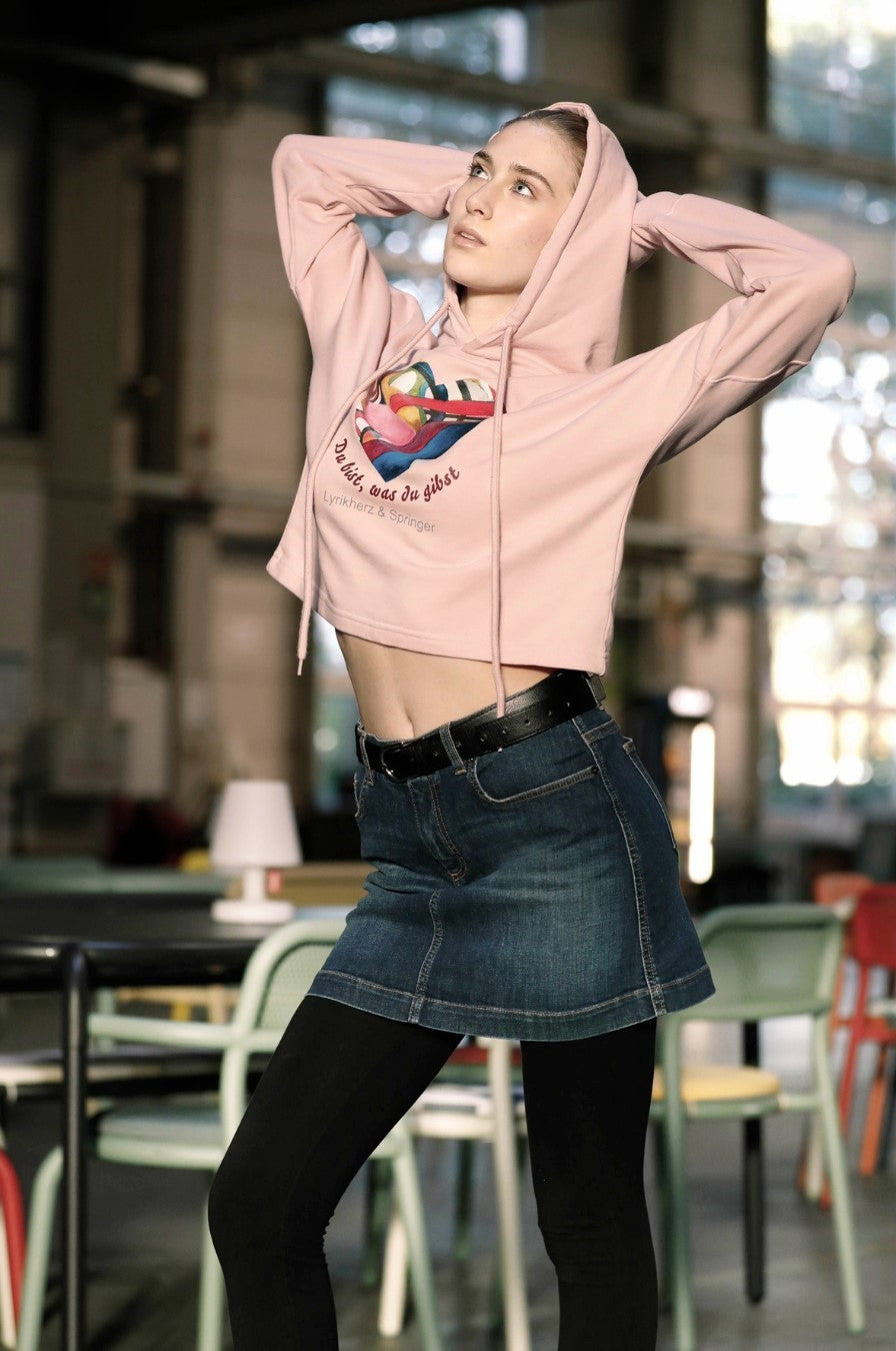 Ein kurz geschnittener, rosafarbener Hoodie mit überschnittenen Schultern. Extralange Bündchen mit Daumenloch und eine bedruckte Kunstgrafik auf der Vorderseite. Mode von Soulfashion Lyrikherz & Springer, erhältlich bei FEMARTSPACE.