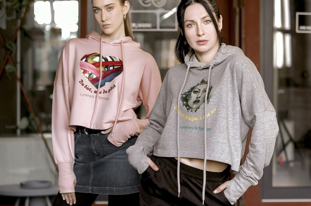 Ein kurz geschnittener, rosafarbener Hoodie mit überschnittenen Schultern. Extralange Bündchen mit Daumenloch und eine bedruckte Kunstgrafik auf der Vorderseite. Mode von Soulfashion Lyrikherz & Springer, erhältlich bei FEMARTSPACE.