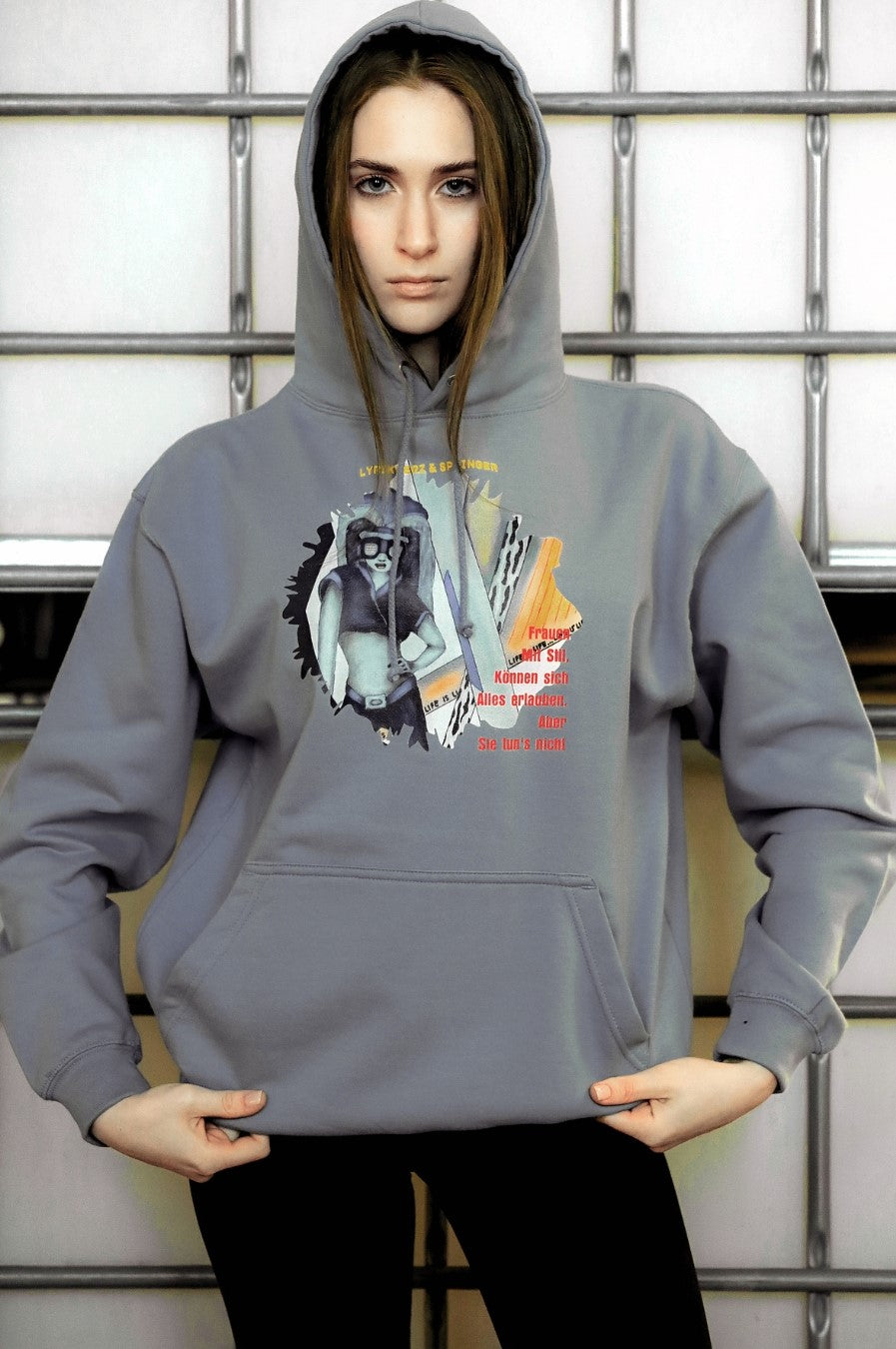 Ein nebelblauer Unisex-Hoodie mit einer Kängurutasche und Kapuze. Auf der Vorderseite ist ein Kunstwerk aufgedruckt: Es zeigt eine stilisierte Frau mit Brille in Collagen-Optik, begleitet von Text in Gelb und Rot. Fashionmode von Soulfashion Lyrikherz & Springer. Erhältlich in der Online-Galerie von FEMARTSPACE.