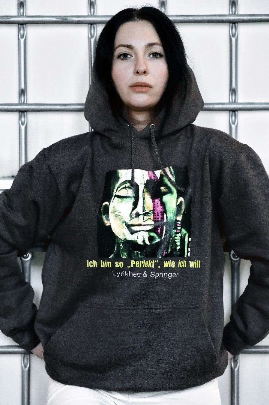 Ein anthrazitfarbener Unisex-Hoodie mit Kängurutasche und Kapuze. Auf der Vorderseite ist ein Kunstwerk gedruckt: Es zeigt ein abstrakt gestaltetes Gesicht, das halb modern, halb klassisch wirkt. Darunter steht der gelbe Schriftzug „Ich bin so 'PERFEKT' wie ich will“, ergänzt durch den Text Lyrikherz & Springer. Erhältlich bei FEMARTSPACE.