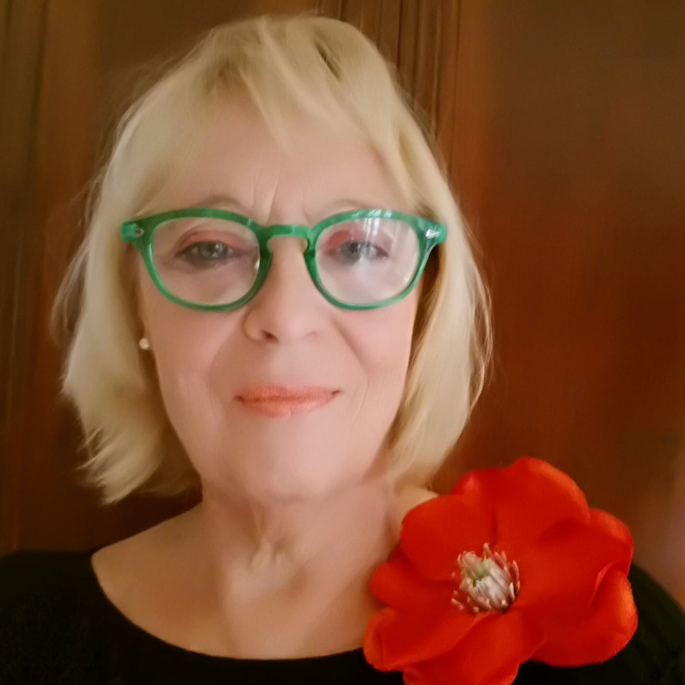 Porträt von Angelika Wesener: Die Künstlerin mit blondem Haar und grüner Brille trägt eine rote, große Ansteckblume an einem schwarzen Oberteil.