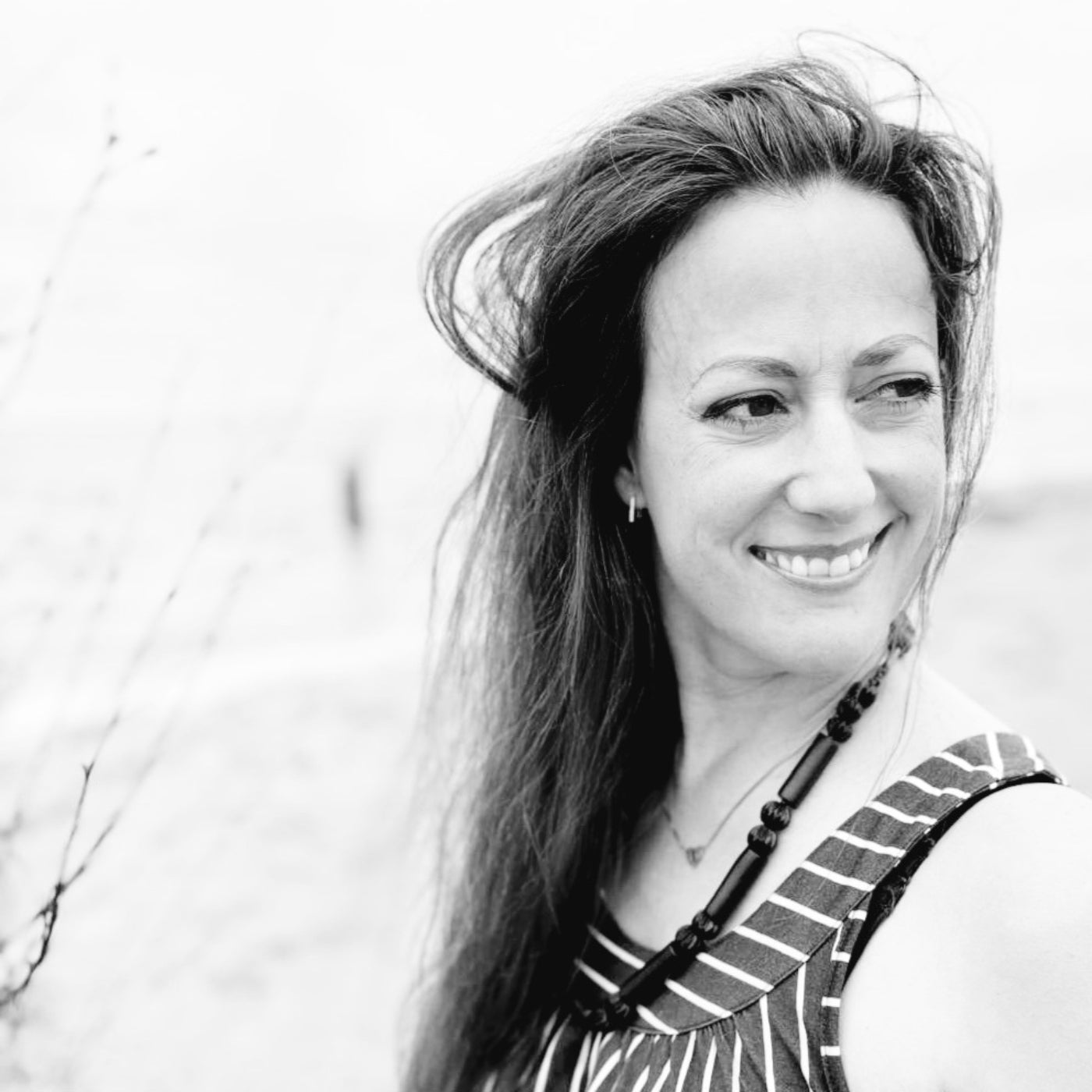 Porträt der Künstlerin Melanie Zürn. Sie steht draußen, blickt lächelnd nach rechts aus dem Bild. Ihr langes Haar weht leicht im Wind. Sie trägt ein gestreiftes ärmelloses Oberteil und eine dunkle Halskette. Das Foto ist in Schwarz-Weiß gehalten.