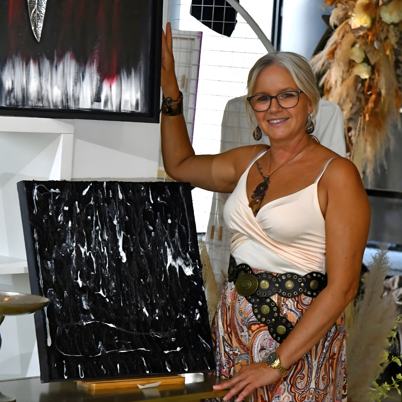 Porträt der Künstlerin Sandra Konrad, die lächelnd neben zwei ihrer abstrakten Kunstwerke steht. Sie hat graue Haare, trägt eine Brille und ist in einem hellen Oberteil und einem gemusterten Rock zu sehen. 