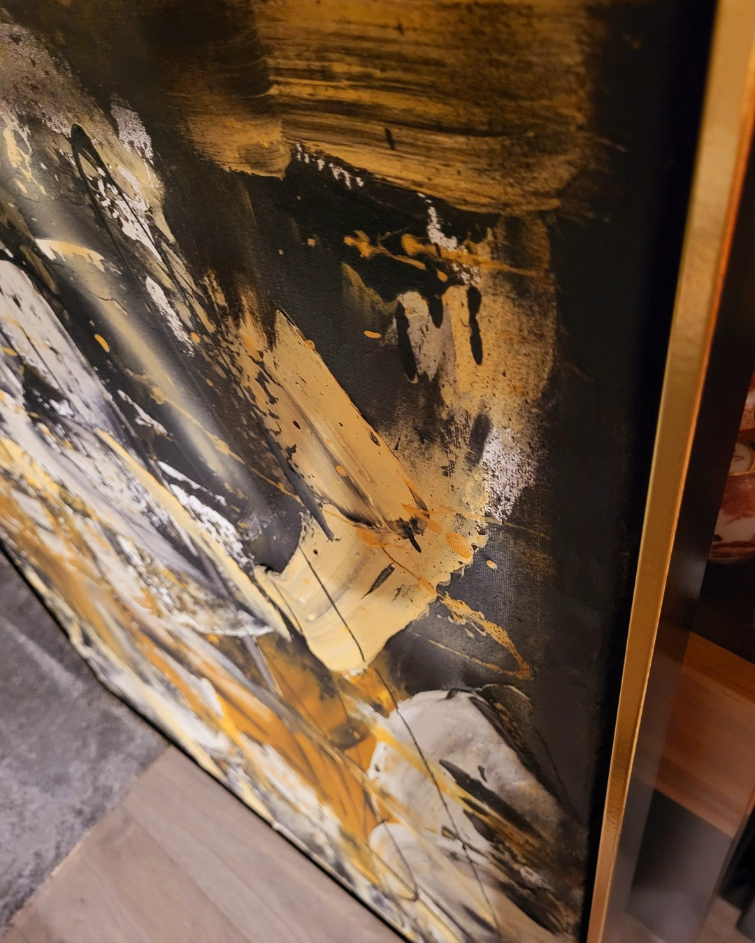 Abstraktes Acrylgemälde "Cocky" von Sandra Conrad, hochformatig, mit Struktur und im schwarz-goldenen Rahmen. Die Komposition besteht aus breiten, gestischen Pinselstrichen in Gold/Ocker, Schwarz und Grau. Freie, schwarze Kritzeleien liegen auf der Farbschicht.