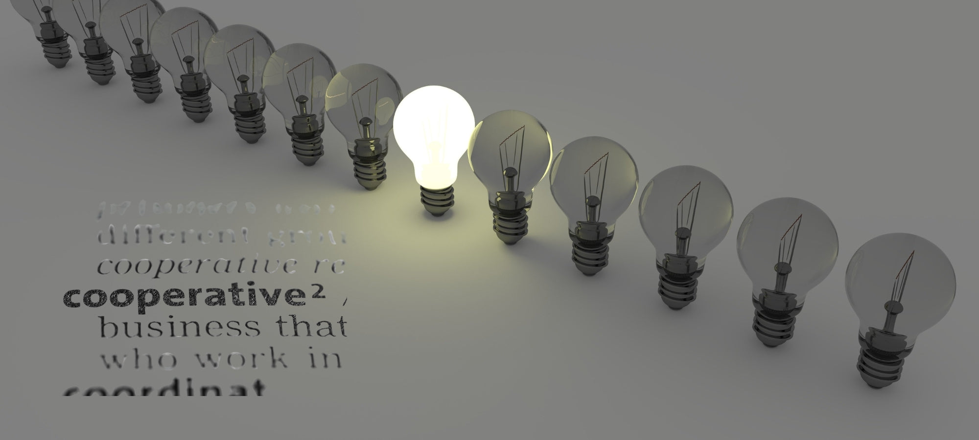 Mehrere Glühbirnen stehen in einer Reihe auf grauem Untergrund. Eine einzelne Birne in der Mitte leuchtet hell, während die übrigen dunkel bleiben. Darunter sind teilweise unscharf englische Wörter wie ‚cooperative‘ und ‚business‘ zu erkennen