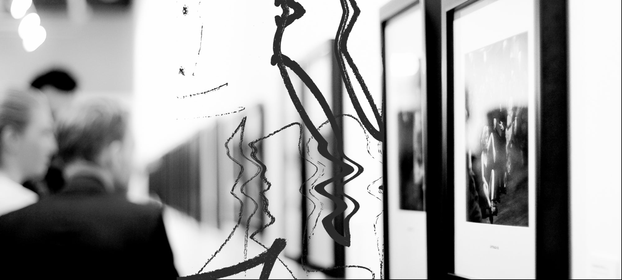Schwarz-weiß Fotografie einer Galerieansicht. Unscharfe Besucher:innen betrachten eine Reihe gerahmter Fotografien an der Wand. Im Vordergrund ziehen sich schwarze, frei geschwungene Linien über die Glasfläche und bilden ein grafisches Muster, das mit den klaren Bildrahmen kontrastiert.