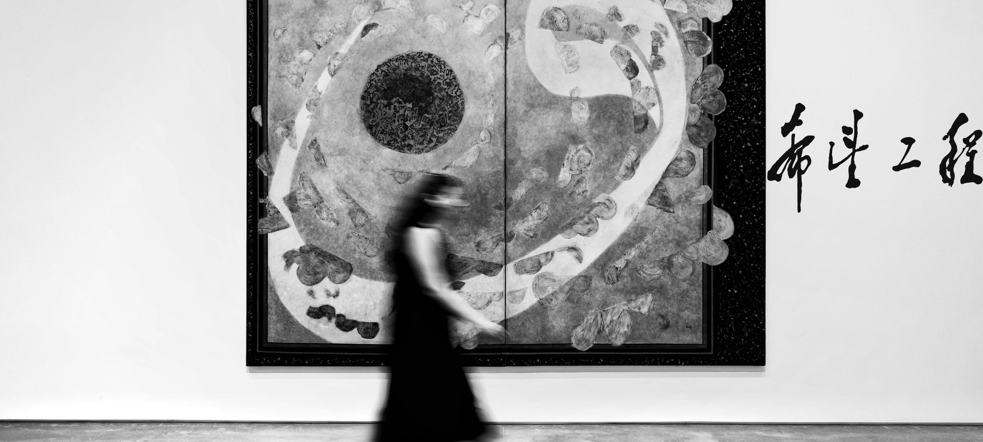 Schwarz-weiß Aufnahme eines großen abstrakten Gemäldes in einer Galerie. Im Vordergrund geht eine Person in fließendem dunklen Kleid unscharf am Bild vorbei. Das Gemälde zeigt kreisende Formen und eine zentrale dunkle Scheibe, rechts daneben schwarze kalligrafische Schriftzeichen auf weißer Wand.
