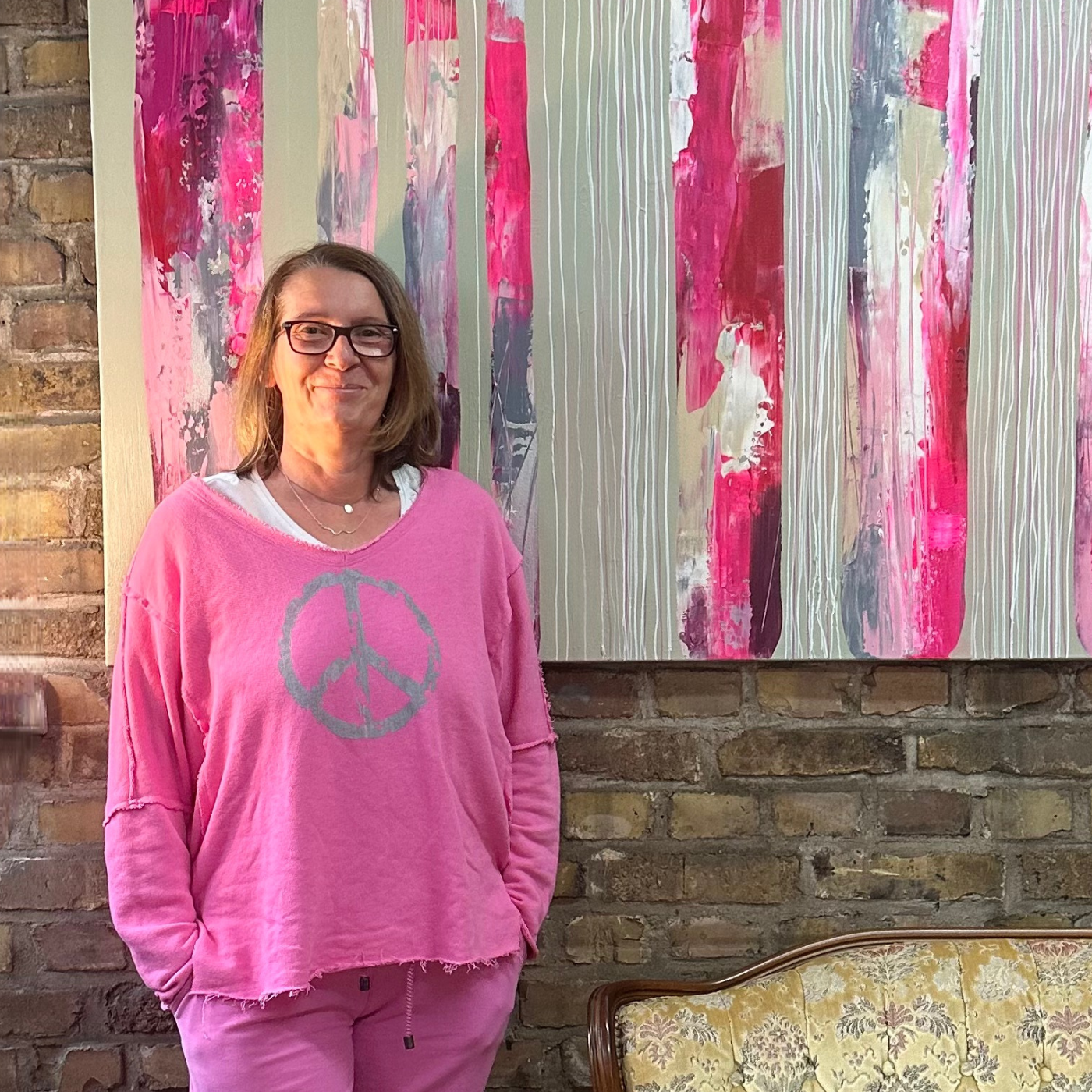 Porträt der Künstlerin Sabine Hoch-Himmighofen, die lächelnd vor einem ihrer abstrakten Gemälde steht. Das Bild zeigt breite vertikale Streifen in leuchtendem Pink, Rot, Grau und Weiß. Die Künstlerin trägt einen pinkfarbenen Pullover mit einem grauen Peace-Zeichen und steht vor einer Ziegelsteinwand.