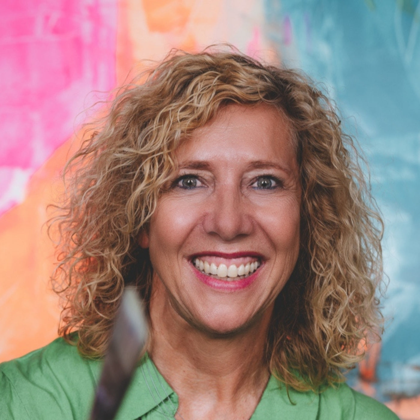 Porträt der lächelnden Künstlerin Susann Versteegen mit lockigen blonden Haaren und einem grünen Hemd. Sie hält einen Pinsel im Vordergrund und ist vor einem farbenfrohen, abstrakt gemalten Hintergrund in Orange, Pink und Türkis zu sehen.
