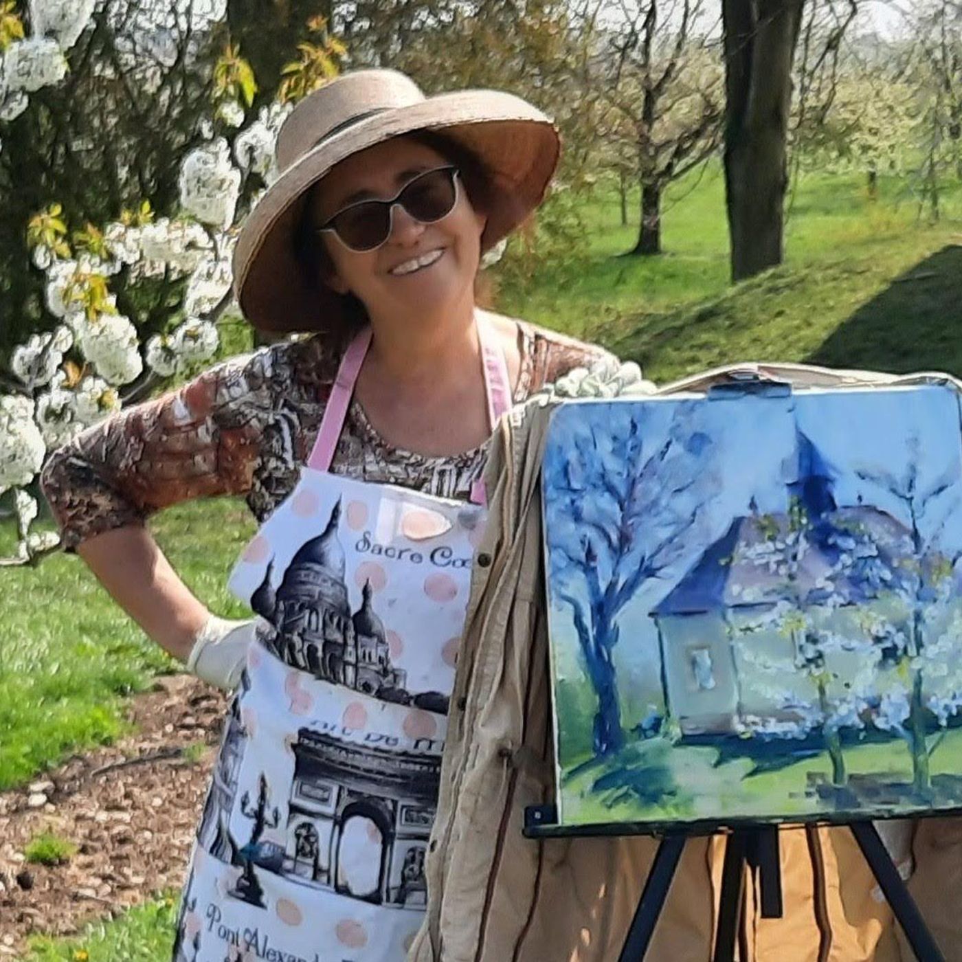 Künstlerin Vera Stein lächelnd in einem Sonnenhut neben einer Staffelei, die ein impressionistisches Frühlingsgemälde zeigt.
