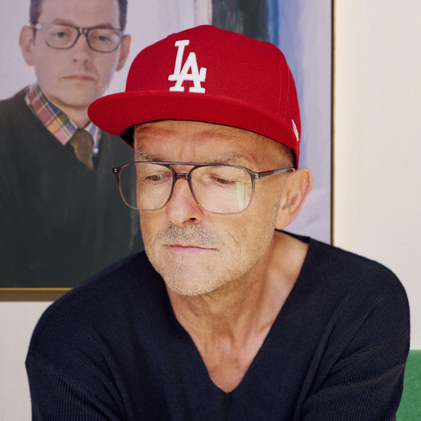 Profilbild des Künstlers nostrokes für die Online-Galerie FEMARTSPACE. Das Bild zeigt ihn in entspannter Pose mit roter Mütze, während im Hintergrund eines seiner typischen, malerischen Porträts auf Leinwand zu sehen ist.